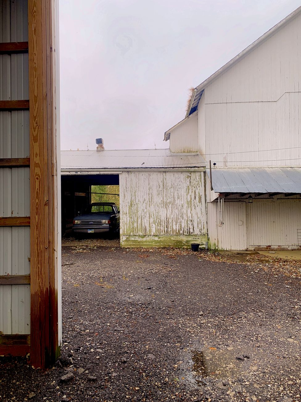 An aged barn with a black car parked inside, captured on a cloudy day in a rural setting.